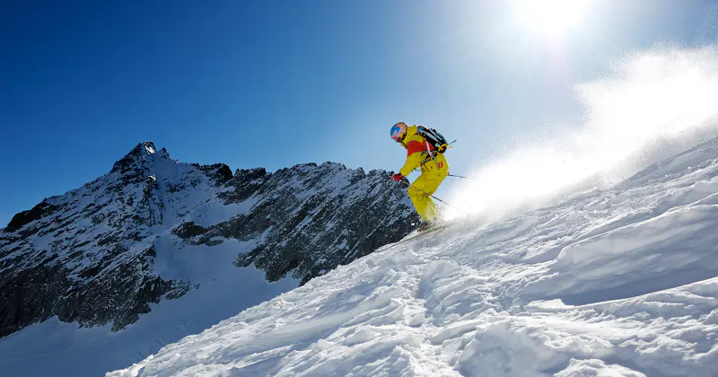 Een skiër in felgele kleding daalt onder een strakblauwe hemel een besneeuwde berghelling af, terwijl het zonlicht boven de grillige bergtoppen op de achtergrond schijnt.